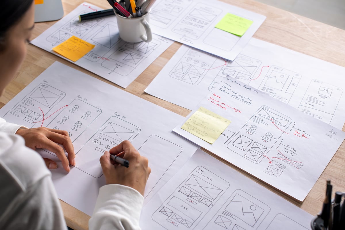 UX UI designer working on wireframes for a website and mobile app prototype, surrounded by sketches of user-flow and design tools